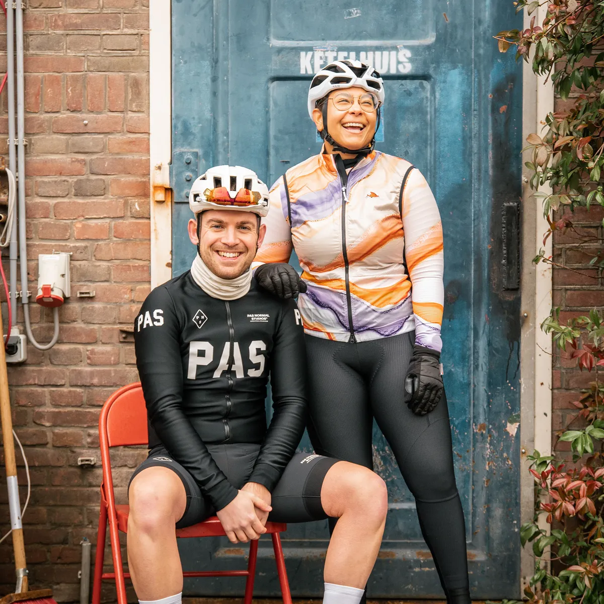 Twee wielrenners in fietskleding poseren lachend voor een blauwe deur