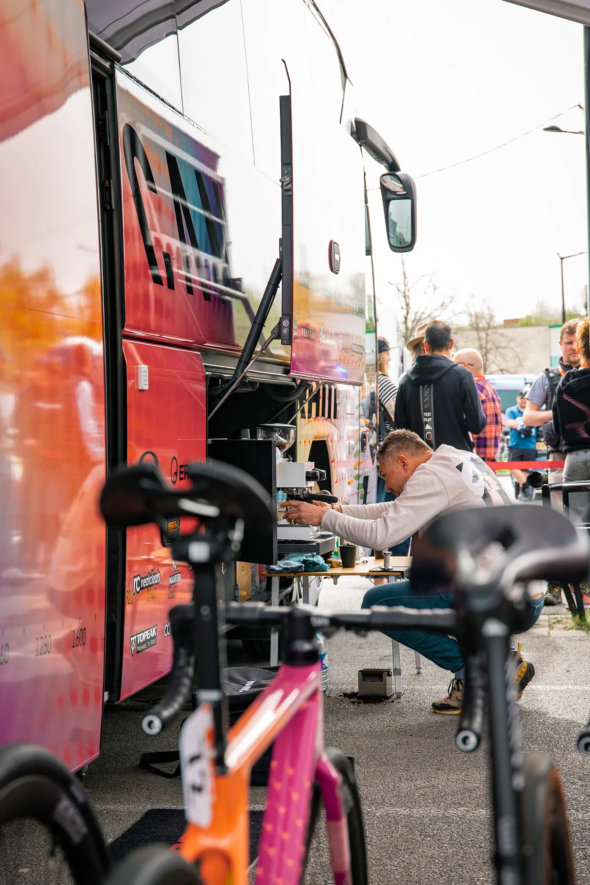 Barista zet espresso naast de teambus, renners op de achtergrond